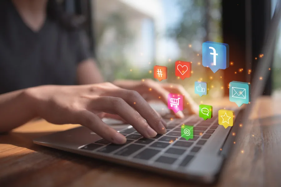 Χέρια που πληκτρολογούν σε laptop, με εικονίδια social media να αιωρούνται πάνω από το πληκτρολόγιο